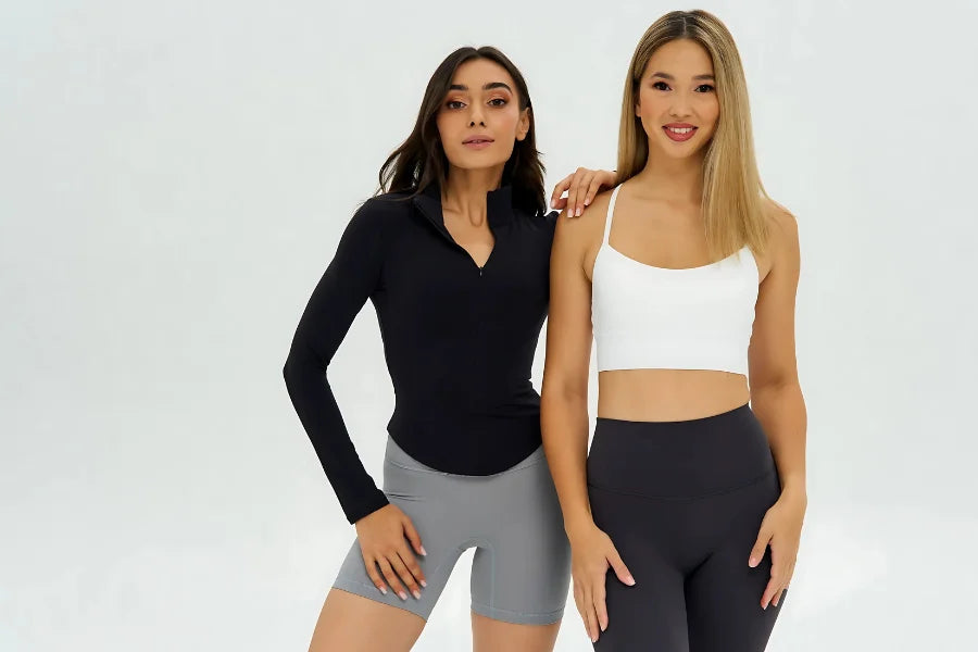 Two models wearing DUNIQ activewear. Black long sleeve on grey bike shorts, and white one-strap sports bra on black leggings.